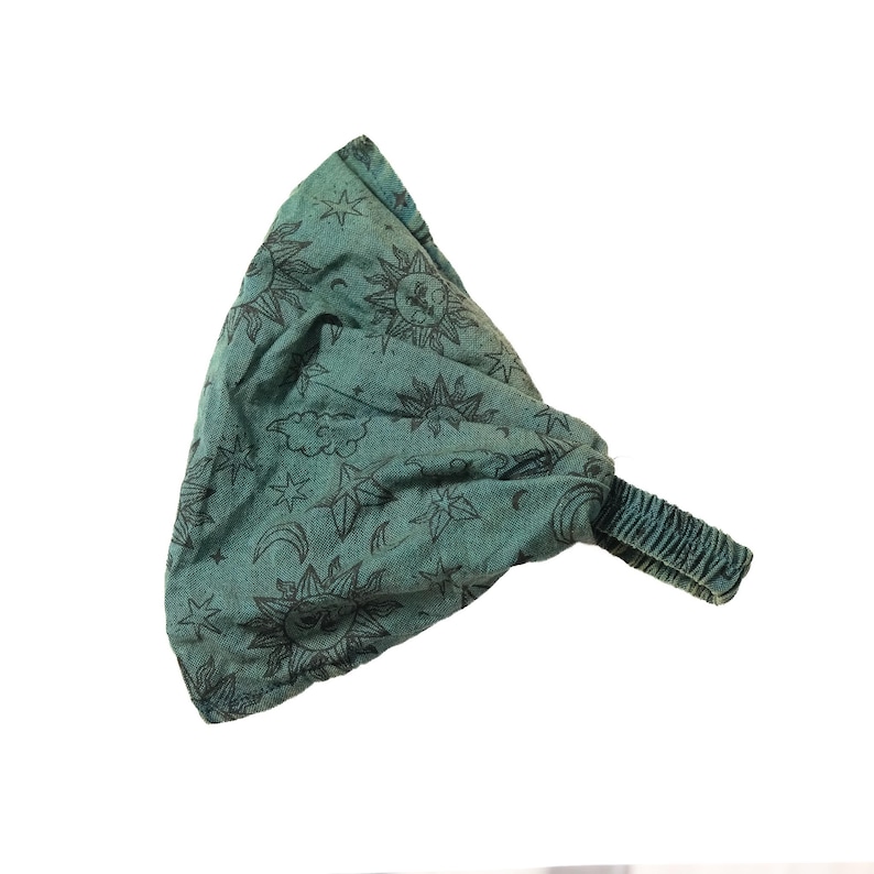 May include: A teal fabric headband featuring a repeating black print of suns, stars, and crescent moons. The accessory has a gathered, wide top section and a narrow, elasticated band at the back to provide a secure fit for the wearer.
