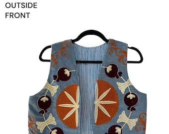Op de afbeelding: Een lichtblauw vest met geborduurde oranje citrusvruchten, donkerpaarse granaatappelachtige vormen en crèmekleurige strikmotieven. De tekst RVHJ 1 OUTSIDE FRONT is zichtbaar in de linkerbovenhoek van de afbeelding.