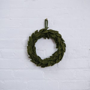 May include: A green felt wreath with a simple design, hanging on a white brick wall.