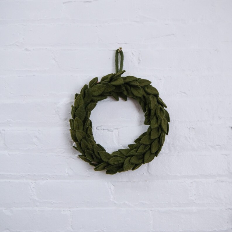 May include: A green felt wreath with a simple design, hanging on a white brick wall.
