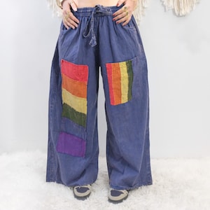 Puede incluir: Pantalones azules de pierna ancha con cintura elástica de cordón y grandes bolsillos rectangulares de parche en la parte delantera. Cada bolsillo muestra un patrón de rayas verticales en segmentos de tela roja, naranja, amarilla, verde y morada.