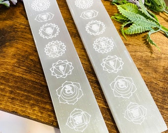 May include: Two white selenite charging wands with seven chakra symbols etched on each. The wands are on a wooden surface.