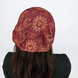 May include: A deep red fabric cap featuring a repeating gold-toned print of suns, crescent moons, stars, and clouds. The head covering has a rounded, slouchy silhouette that fits over the head, displaying a celestial pattern throughout.
