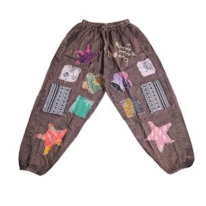 May include: Brown trousers with an elasticated waistband and drawstring, featuring various colourful fabric patches in star, rectangular, and square shapes. The patches include floral, striped, and geometric patterns sewn onto the dark brown fabric.