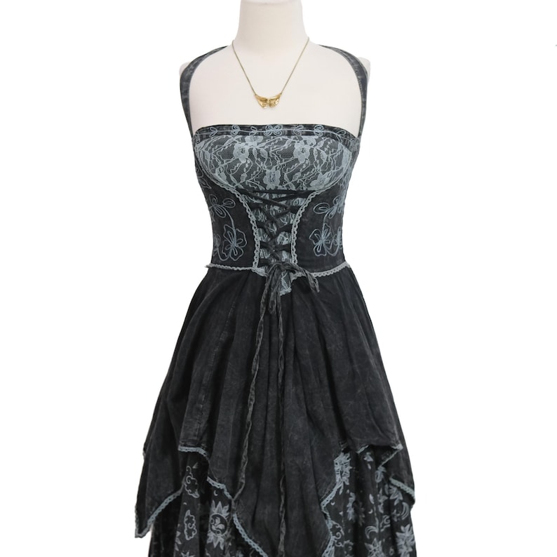 May include: A black and grey halter dress featuring a lace-up corset-style bodice with floral embroidery. The dress has a layered, uneven hemline and a fitted waist, displayed on a mannequin with a gold-toned necklace featuring a butterfly pendant.