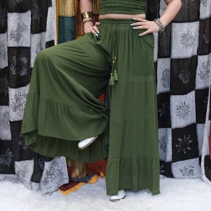 May include: Olive green wide-leg pants featuring a high-waisted design with an elastic waistband, a drawstring closure with tassel ends, two side pockets, and a tiered ruffled hemline that creates a voluminous, flowing silhouette.