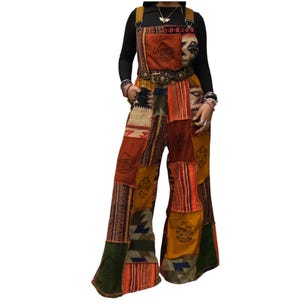 May include: A pair of patchwork overalls featuring a variety of earth-toned fabric panels in shades of brown, orange, and green. The garment includes a front chest pocket with a mushroom graphic, wide legs, and adjustable shoulder straps.