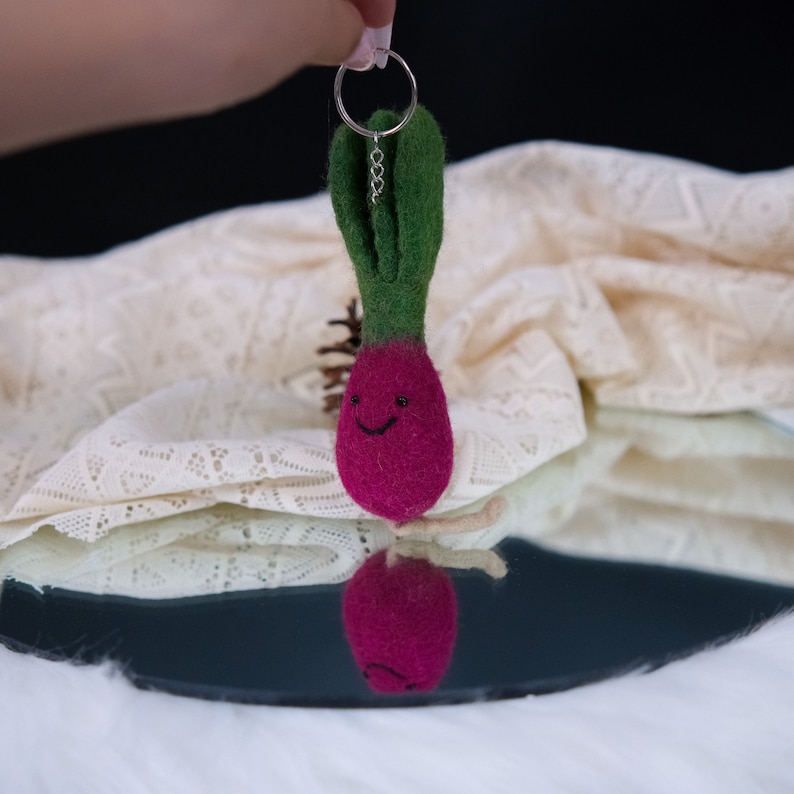 May include: A felt keychain shaped like a radish with a green stem and a pink body. The radish has a smiling face.