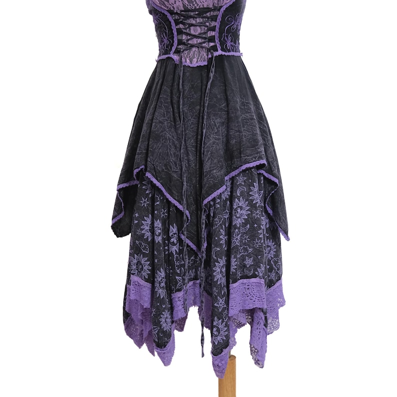 May include: A sleeveless dress featuring a black and purple color scheme with a lace-up corset bodice. The skirt has an asymmetrical handkerchief hemline, a celestial sun and moon print, and purple lace trim along the bottom edges.