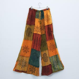 Könnte beinhalten: Weit geschnittene Hose mit einem Patchwork-Design aus rechteckigen Stoffbahnen in Orange, Gelb, Rot und Grün. Der Stoff ist mit schwarzen grafischen Linien bedruckt, die Sonnen, Augen und Hamsa-Hand-Symbole auf dem gesamten Kleidungsstück zeigen.