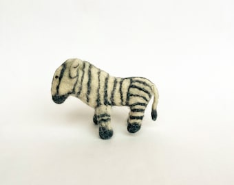 May include: A small, hand-felted zebra toy made of white and black wool. The zebra is standing on a white surface.