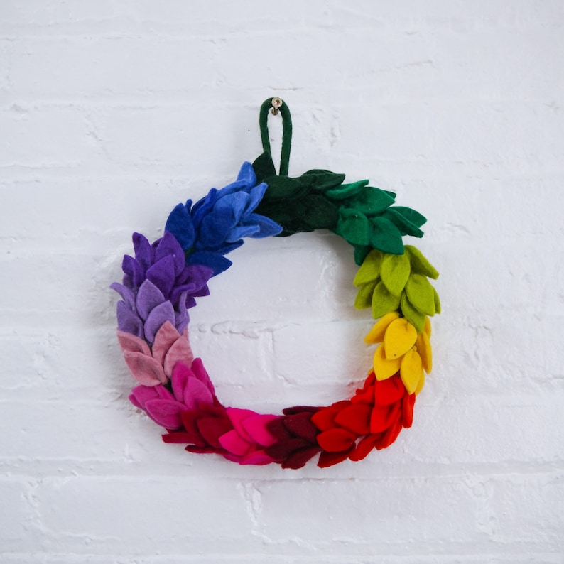 May include: A rainbow felt wreath with a green felt center and a brown hanging loop. The wreath is made of felt leaves in shades of blue, purple, pink, red, orange, yellow, and green.