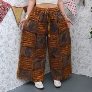 May include: Brown and orange patchwork pants with a wide leg design. The pants feature embroidered floral, moon, and star patterns. The waist has a drawstring. The pants are made of a soft, textured fabric.