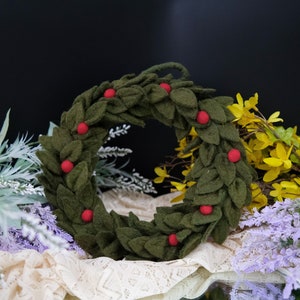 May include: A green felt wreath with red felt berries. The wreath is sitting on a white lace tablecloth with purple and yellow flowers in the background.