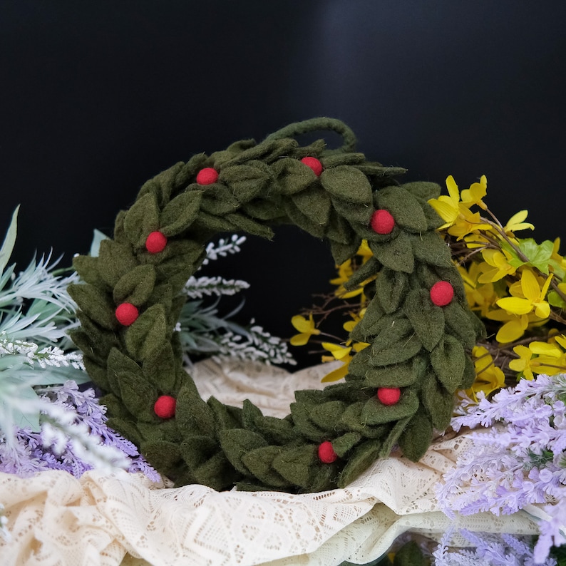 May include: A green felt wreath with red felt berries. The wreath is sitting on a white lace tablecloth with purple and yellow flowers in the background.
