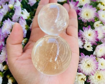 Op de afbeelding: Twee heldere kwartskristallen bollen, een met een gouden tint, rustend in een hand tegen een achtergrond van paarse en witte bloemen.