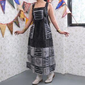 May include: A black and gray denim dress with a square neckline and wide straps. The dress features multiple pockets and a patchwork design with floral and star patterns. The model is wearing white boots and gold bracelets.