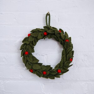May include: A green felt wreath with red felt berries. The wreath is hanging on a white brick wall.