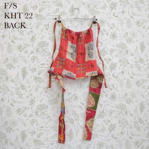 May include: A red, green, and beige patchwork halter top featuring floral and geometric patterns with visible stitching. The garment includes long fabric ties and is labeled with the text F/S KHT 22 BACK. It is displayed on a metal hanger.
