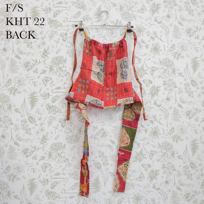 May include: A red, green, and beige patchwork halter top featuring floral and geometric patterns with visible stitching. The garment includes long fabric ties and is labeled with the text F/S KHT 22 BACK. It is displayed on a metal hanger.