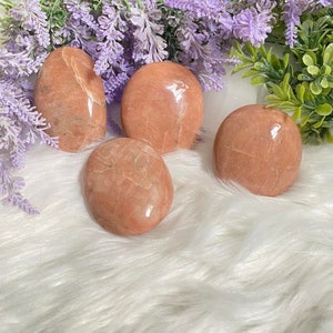 Può includere: Quattro pietre lucide color pesca con finitura liscia e lucida. Le pietre variano nella forma, alcune ovali e altre più rotonde. Lo sfondo comprende lavanda viola e fogliame verde, su una superficie bianca e soffice.