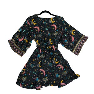 May include: A black short-length dress featuring a celestial print with yellow and pink crescent moons, colorful stars, and floral motifs. The dress has wide, flared sleeves with a geometric patterned border and a tie-waist belt detail.