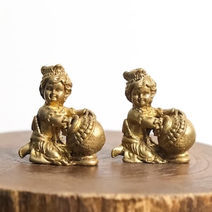 May include: Two identical gold-colored metal figurines depicting a small child kneeling and holding a large, round, textured pot. Each figure features detailed facial expressions, curly hair, and traditional clothing, with a metallic finish.