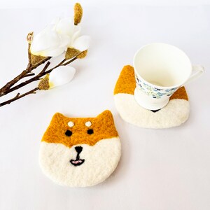 May include: Two felt coasters, one with a brown and white Shiba Inu design and the other with a brown and white design. The coasters are on a white surface.