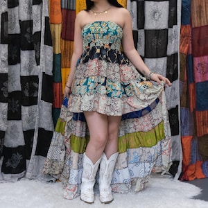 Puede incluir: Un vestido palabra de honor con bajo asimétrico y diseño de patchwork con varios estampados en tonos azul verdoso, negro, blanco y verde lima. La falda tiene una estructura de volantes con el dobladillo trasero más largo y botas vaqueras.