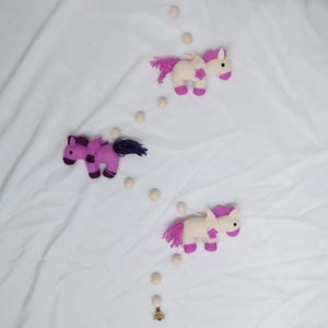 May include: A white, pink, and purple felt unicorn mobile with wooden beads hanging from a string. The mobile features three unicorns with pink and purple details.