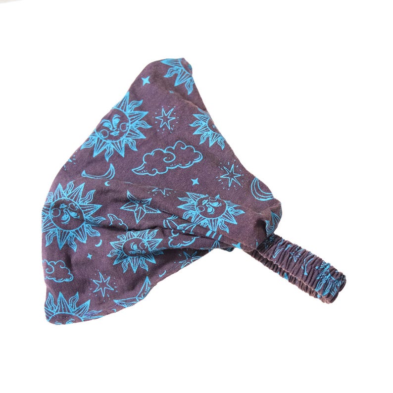 May include: A dark brown fabric headband featuring a repeating pattern of bright blue suns, stars, clouds, and crescent moons. The accessory includes a gathered elastic band section at the back to provide a secure fit for the wearer.