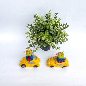 以下が含まれることがあります： 青いフェルトのギフトボックスが載った、黄色のフェルトの玩具の車が2台。車は白い表面にあり、背景には緑色の鉢植えの植物があります。