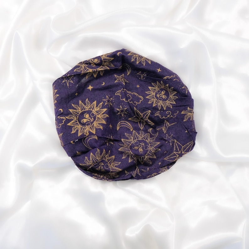 May include: A dark purple fabric head covering featuring a repeating gold-coloured print of suns with faces, crescent moons, stars, and clouds. The circular item has a gathered, rounded shape and is displayed as a single, folded textile accessory.