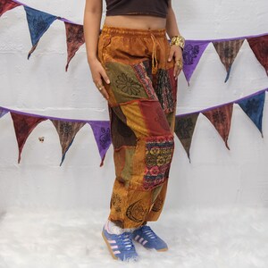 May include: Loose-fitting pants featuring a patchwork design in shades of brown, orange, and red. The fabric displays various black printed motifs, including flowers, moons, and geometric patterns. The pants have an elastic waistband with a drawstring.