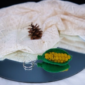 May include: A felt corn on the cob keychain with a green husk and yellow kernels. The keychain has a silver ring attached to it.