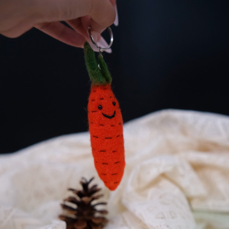 May include: A felt carrot keychain with a smiling face and green leaves. The keychain is attached to a silver key ring.