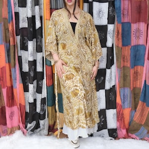 May include: A long, flowing kimono-style robe in a light gold and cream color palette. The garment features an intricate, repeating floral and botanical pattern throughout, with a wide, decorative border along the front opening and wide sleeves.