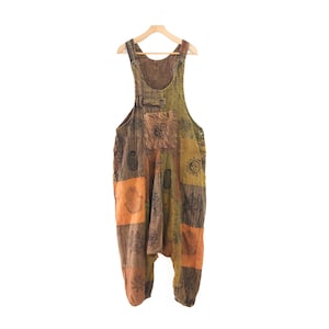 May include: A pair of patchwork overalls in shades of olive green, brown, and orange. The garment features a square chest pocket and a drop-crotch silhouette, decorated with black printed motifs including suns, hamsa hands, and floral patterns.