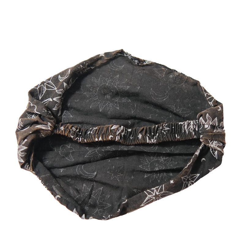 May include: A black fabric headband featuring a white celestial print of suns, moons, and stars. The accessory has a gathered design with an elasticated band at the back to provide a secure fit, showcasing a repeating pattern across the entire surface.
