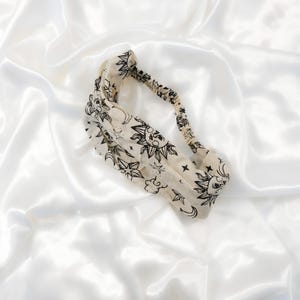 May include: A cream-coloured fabric headband featuring a repeating black ink print of suns, crescent moons, and stars. The headband has a twisted front design and an elasticised back section to provide a secure fit around the head.