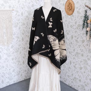 May include: A black shawl with a butterfly pattern is draped over a white dress form. The shawl features a large butterfly design and smaller butterfly accents in white and gray. The edges of the shawl have a fringed trim.