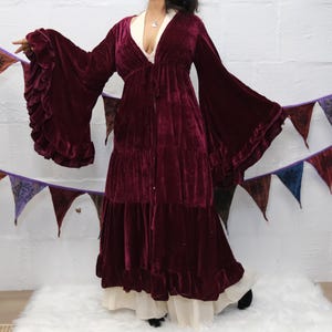 May include: A deep burgundy velvet duster coat featuring long, wide bell sleeves with tiered ruffle cuffs. The garment has a V-neckline, a tie-front closure, and a tiered skirt design that falls to the floor, layered over a cream-coloured garment.