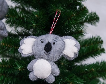 Può includere: Un ornamento di koala in feltro grigio con orecchie bianche e pancia bianca. L'ornamento è appeso a una corda rossa e bianca su un albero di Natale verde.