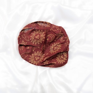 May include: A deep red fabric head covering featuring a repeating gold-toned print of suns with faces, crescent moons, stars, and clouds. The material appears lightweight and slightly sheer, with a gathered, rounded shape and soft folds.