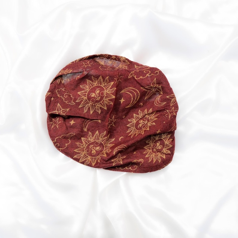 May include: A deep red fabric head covering featuring a repeating gold-toned print of suns with faces, crescent moons, stars, and clouds. The material appears lightweight and slightly sheer, with a gathered, rounded shape and soft folds.