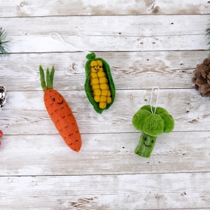 May include: Three felt ornaments shaped like a carrot, corn on the cob, and broccoli. Each ornament has a smiling face and a loop for hanging.