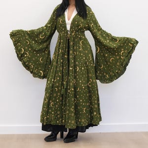 May include: An olive green floor-length duster cardigan featuring a gold celestial print of moons and stars. The garment has long, wide bell sleeves with ruffled edges, a tiered skirt design, and a thin tie closure at the waist.