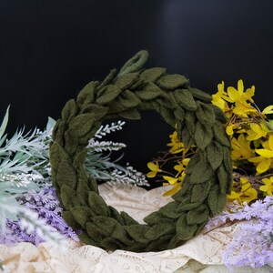 May include: A green felt wreath with a leafy design. The wreath is made of individual felt leaves that are sewn together to form a circle. The wreath is sitting on a white lace fabric.