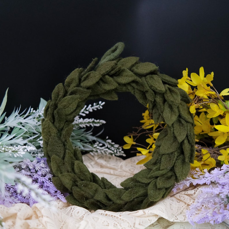 May include: A green felt wreath with a leafy design. The wreath is made of individual felt leaves that are sewn together to form a circle. The wreath is sitting on a white lace fabric.