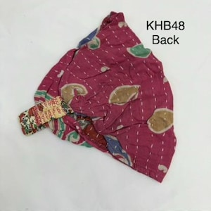 May include: A magenta fabric head wrap featuring a repeating pattern of tan and green shapes with white running stitch details. The item includes an elasticized band with a multi-colored floral print and the text KHB48 Back printed in black.